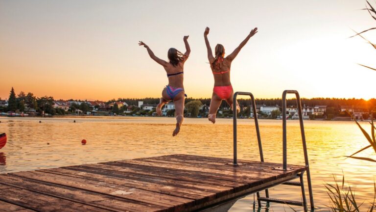 Two women jump off the end of a dock into a lake during summer