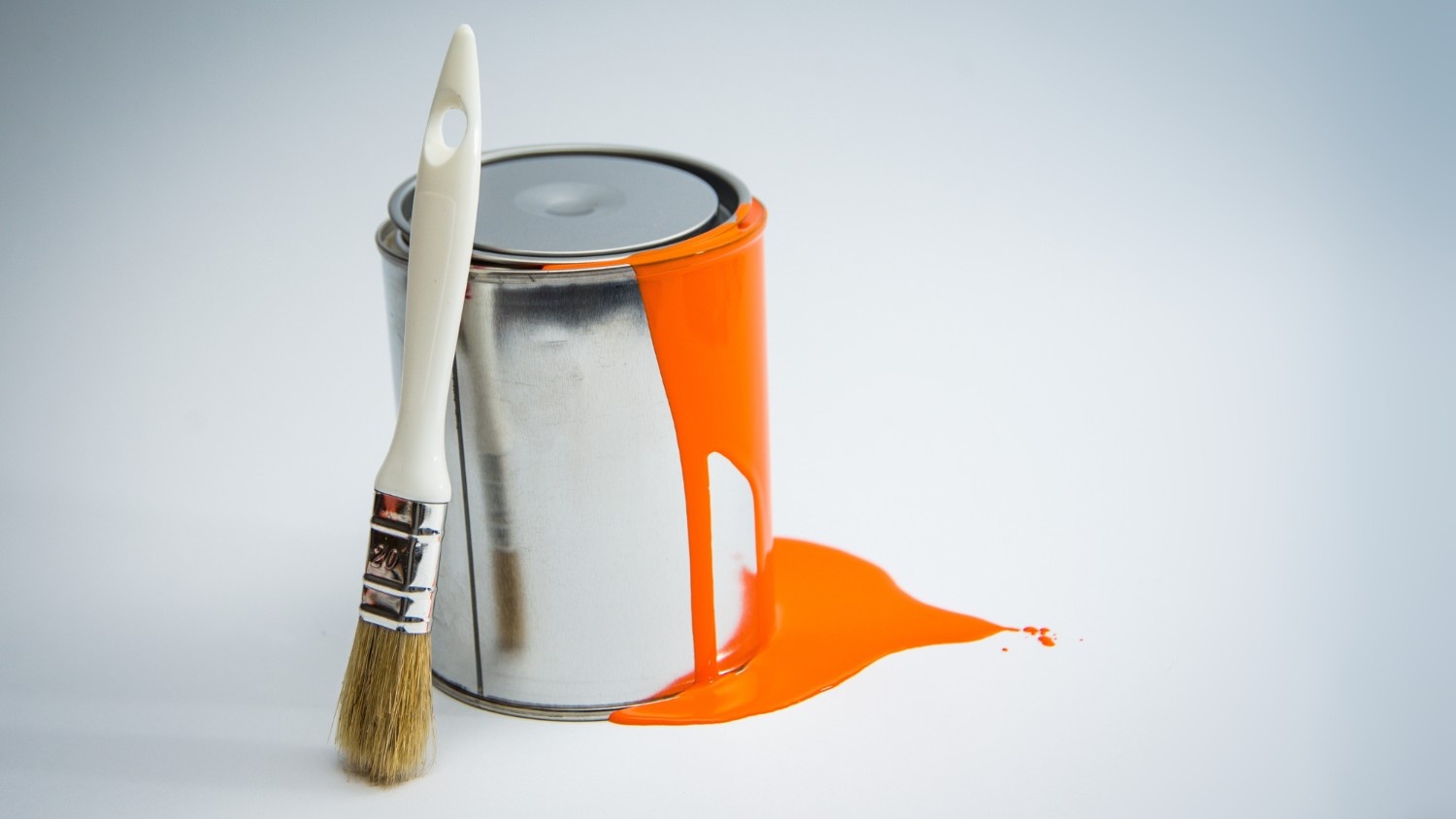 A paint can that is spilling orange paint next to a dry paint brush