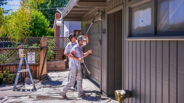 One painter with a roller and another with a sprayer paint the exterior of a home