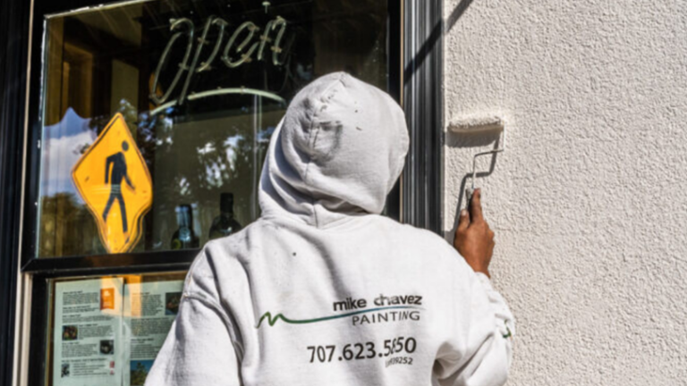 A painter in a “Mike Chavez Painting” sweatshirt paints the exterior of a business