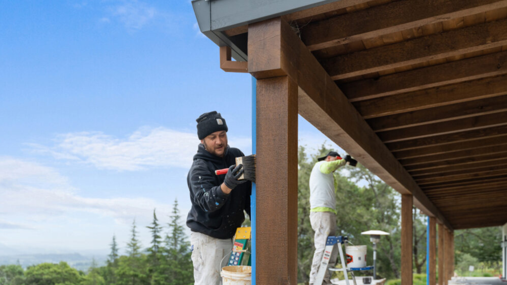 A painter in a beanie and sweatshirt uses a brush on the exterior of a building