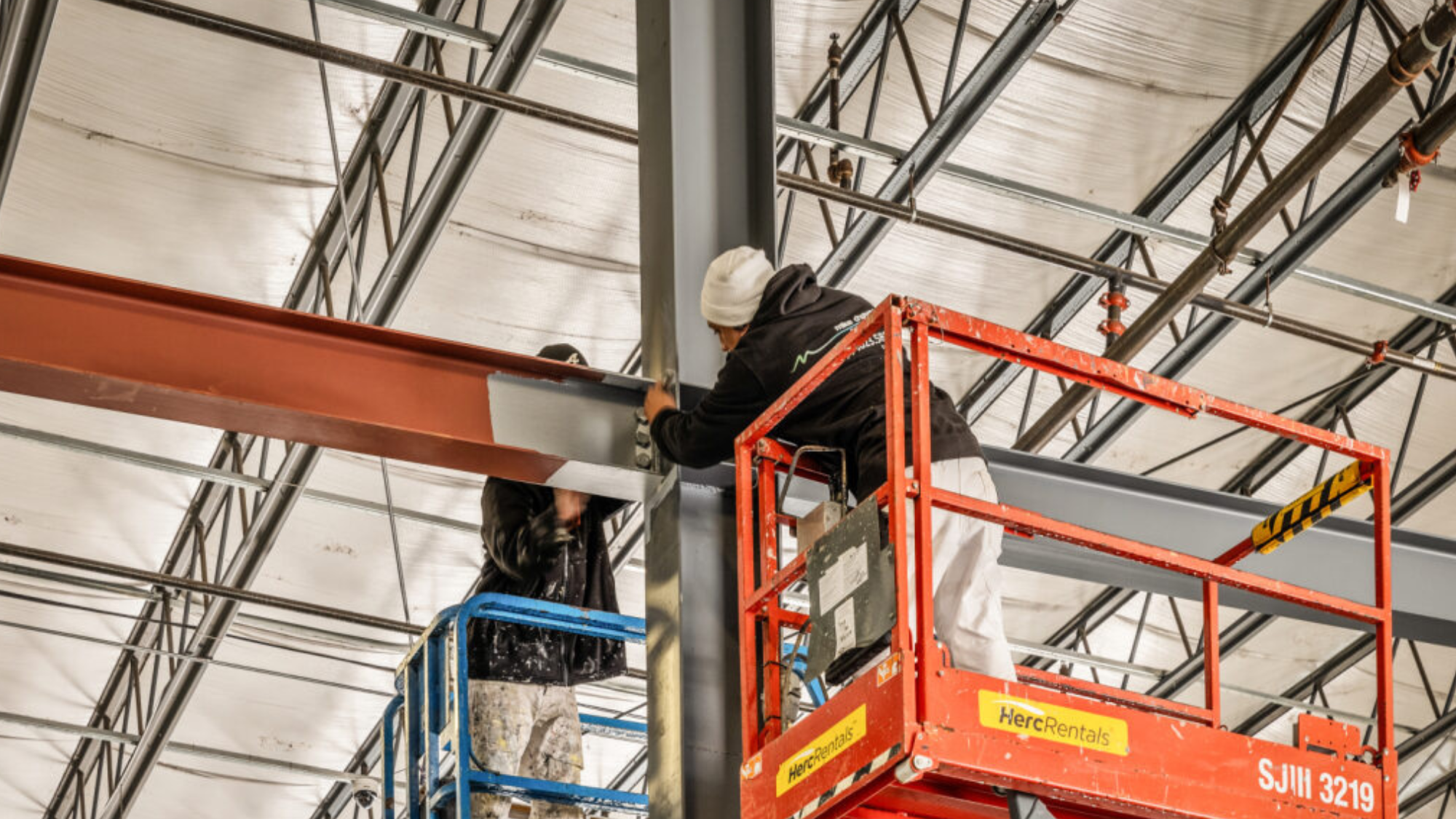 Two painters on lifts paint the metal beams of an industrial building