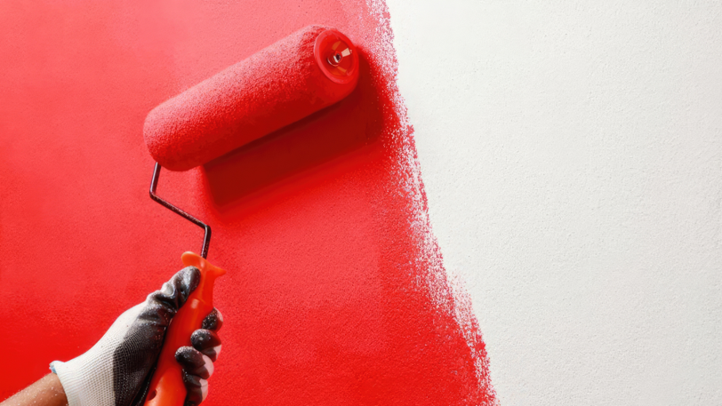 A handheld paint roller being used by a person wearing gloves to paint a wall red.